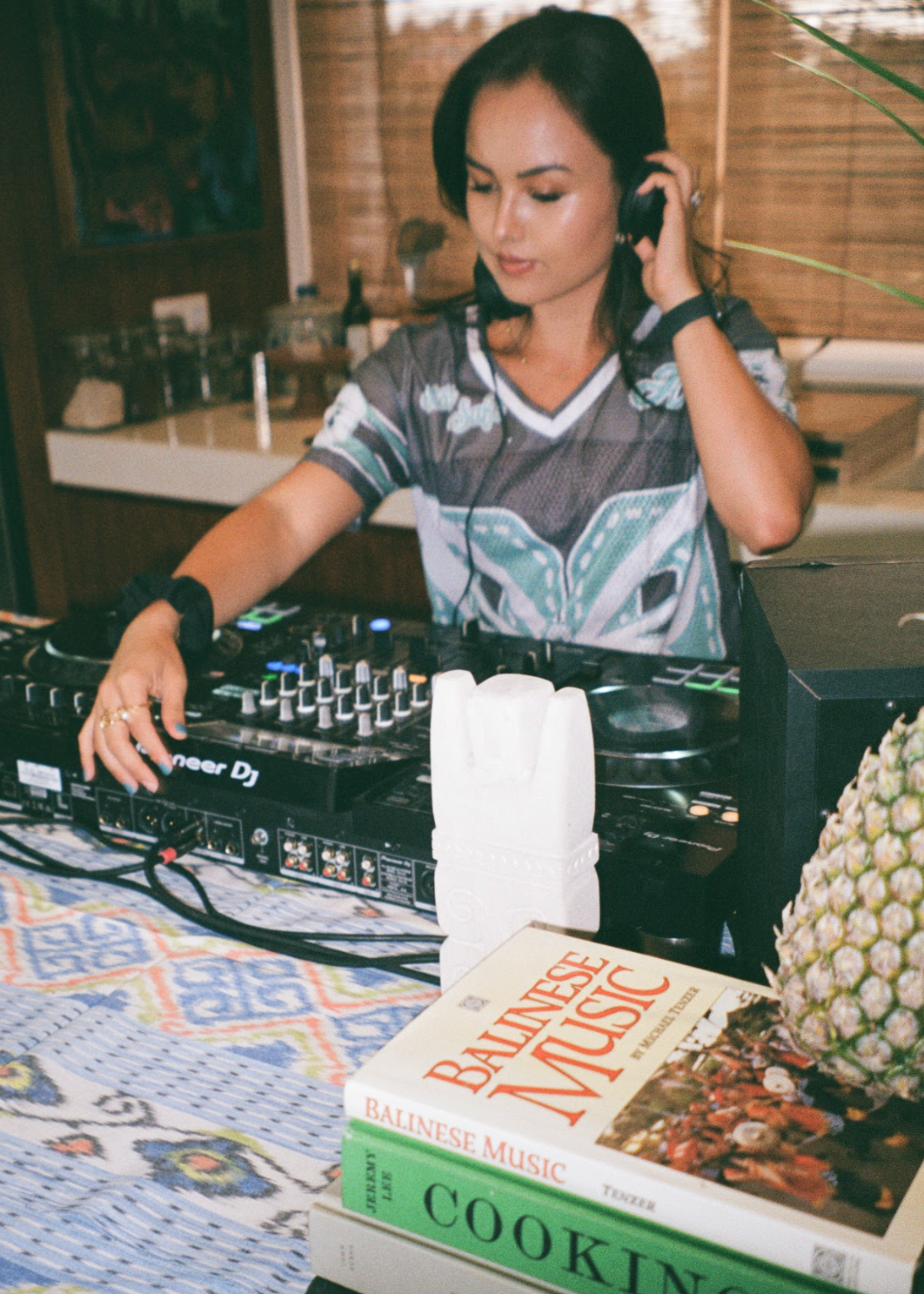 Woman operating DJ equipment with a book titled 'Balinese Magic Cooking' on a table.